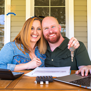An image of a smiling couple in front of their new home, holding keys and surrounded by a stack of paperwork, a calculator, and a laptop