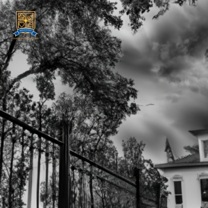 An image of a historic property with unique architectural features, surrounded by mature trees and a wrought iron fence, under a stormy sky